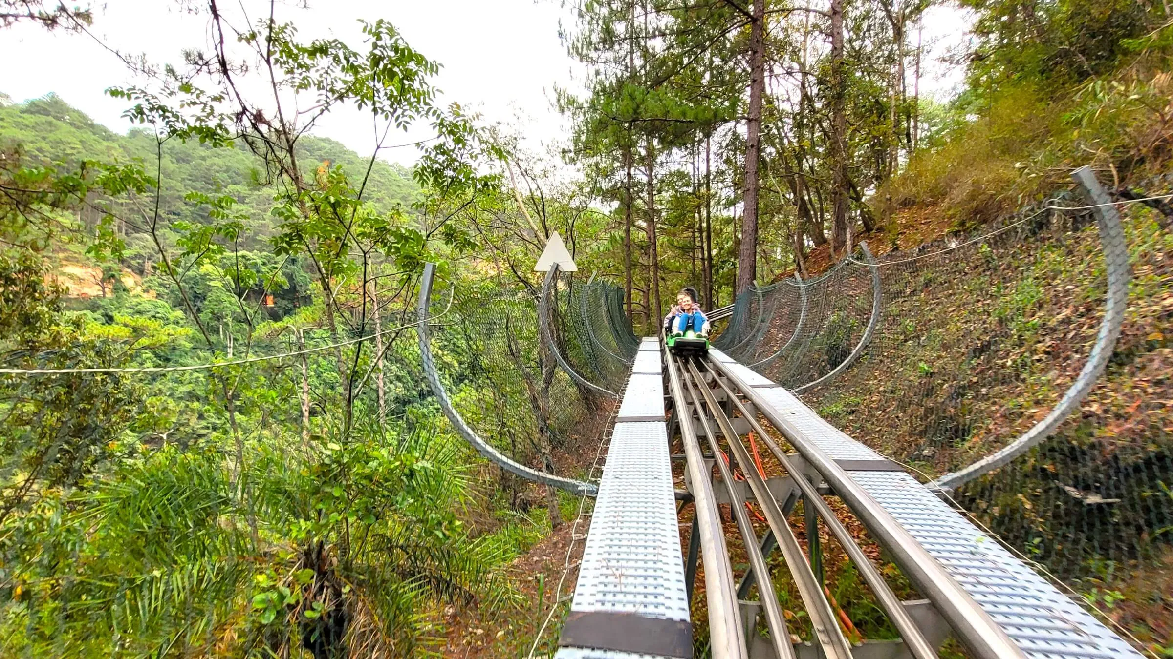 Two smiling riders on a green alpine coaster sled coming down a steel track through the pine forest at Datanla, between curved safety nets