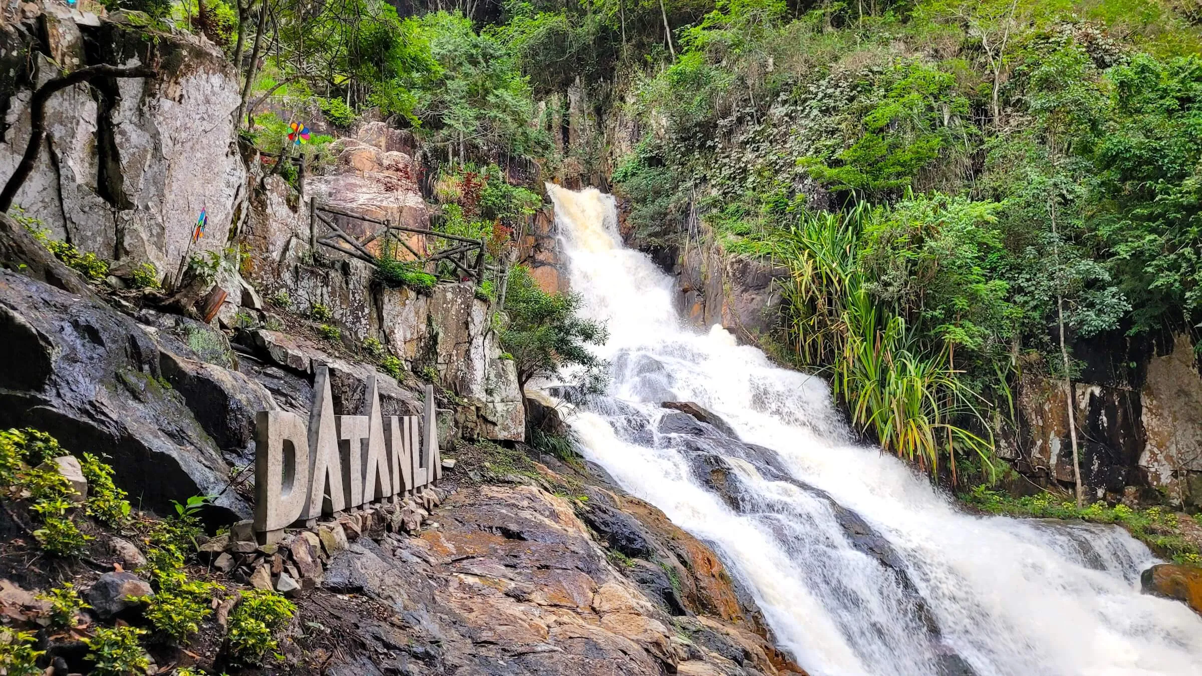 Datanla Waterfall pouring down a rocky slope beside large stone letters spelling DATANLA, with forest and a wooden railing above